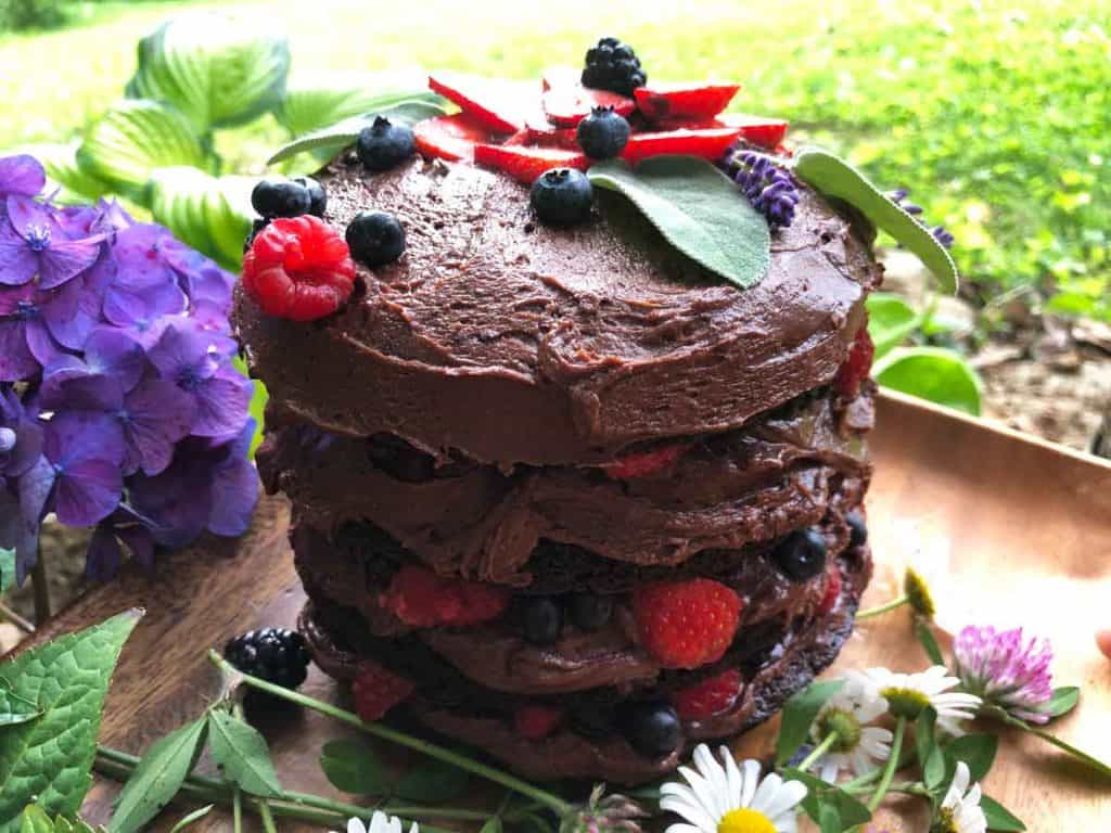 A chocolate cake decorated with fruit is outdoors among purple hydrangea flowers.