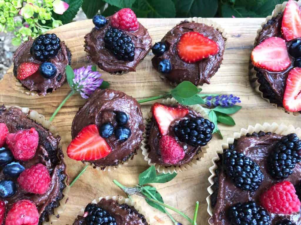 Miniature and full-sized chocolate einkorn cupcakes are decorated with fruit and flowers on a wooden surface.