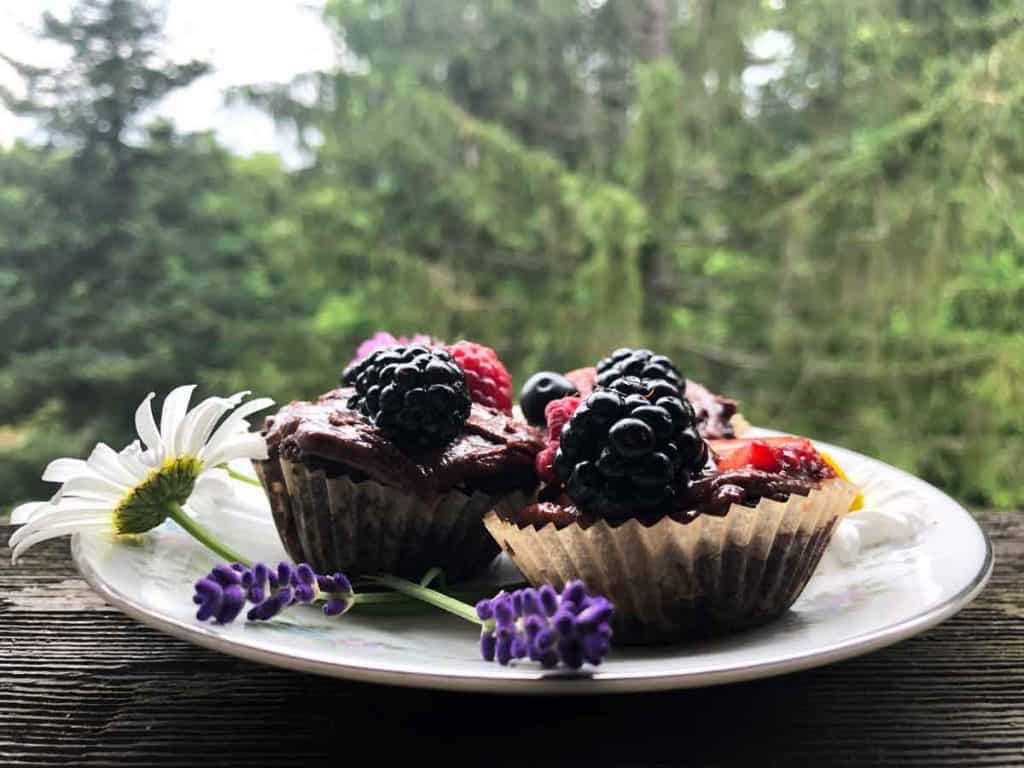Three mini cupcakes are on a dish decorated with flowers in front of evergreen trees.