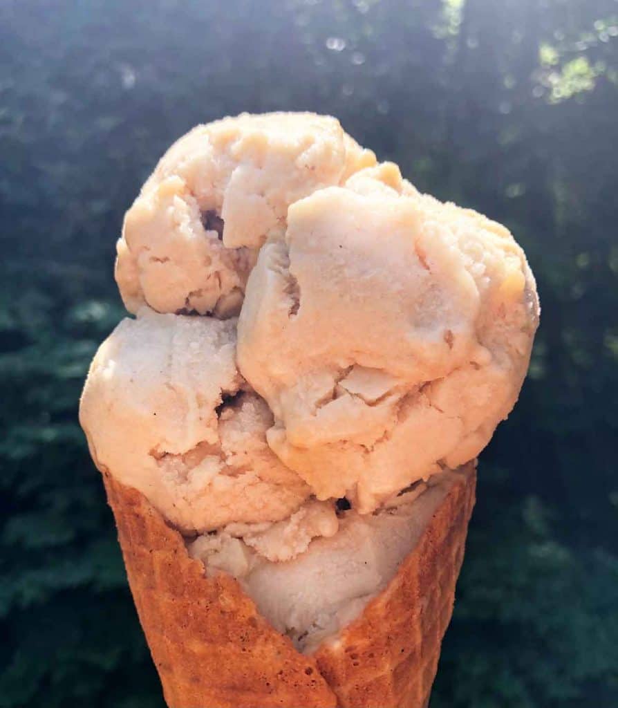 Scoops of vanilla vegan ice cream are in a waffle cone outdoors in the sunshine.