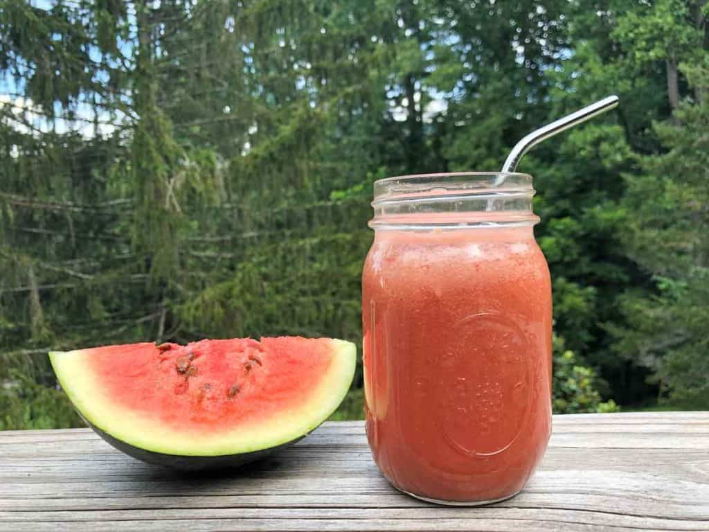 A glass of watermelon juice with a stainless steel straw is beside a slice of watermelon. They are in front of evergreen trees.