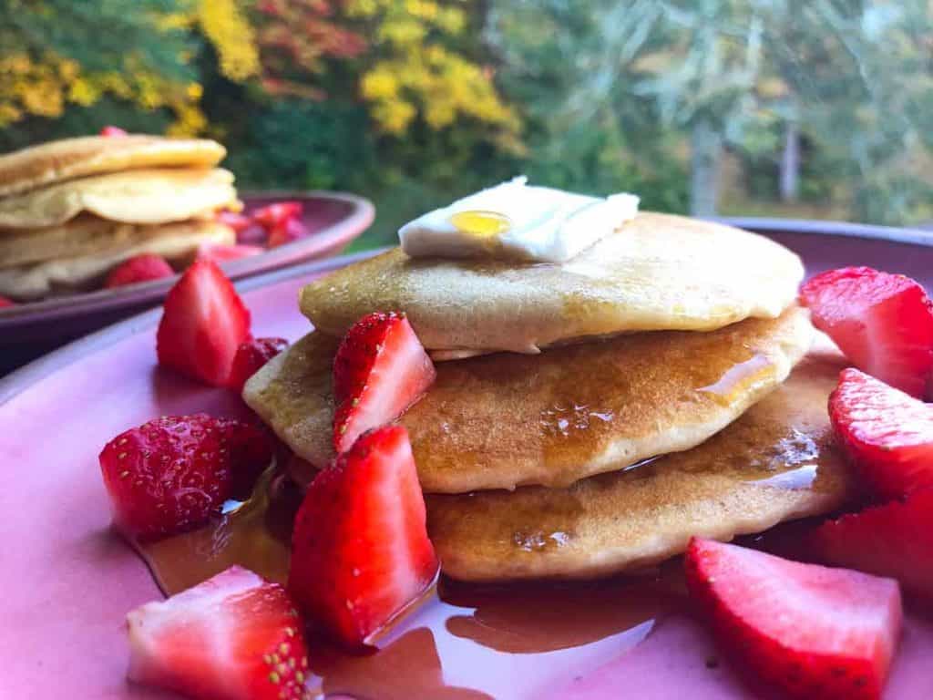 Two pink plates are stacked with einkorn pancakes topped with strawberries and maple syrup. Autumn leaves are in the background.