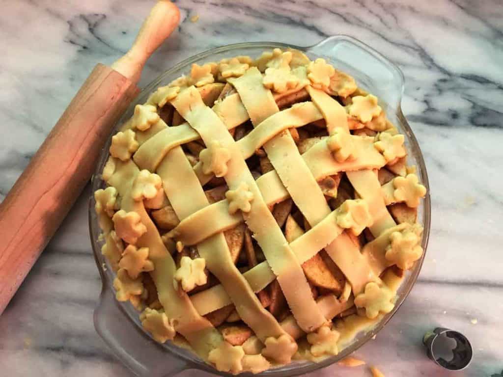 An einkorn apple pie with a lattice crust is beside a wooden rolling pin and flower cookie cutter on a marble countertop.