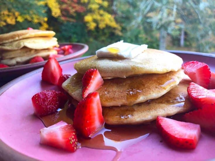 Two plates of pancakes topped with maple syrup and strawberries are on pink plates outdoors in autumn.