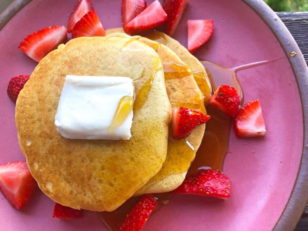 A stack of einkorn pancakes is topped with vegan butter, strawberries, and maple syrup on a pink plate.