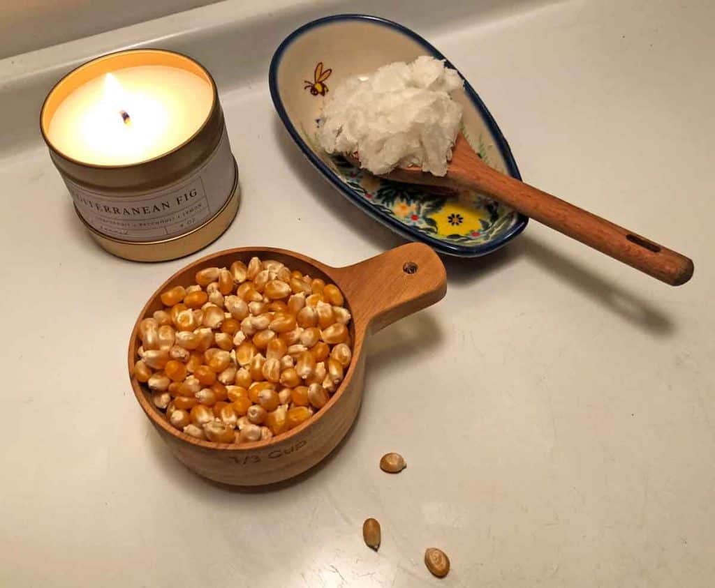 A wooden measuring cup filled with popcorn kernels is beside a wooden measuring spoon full of coconut oil. A lit candle is next to them.