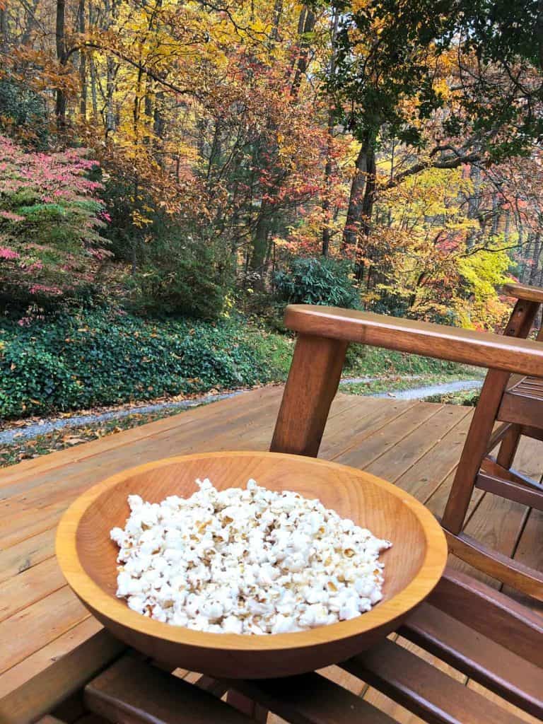 Homemade popcorn is in a wooden bowl resting on a rocking chair outdoors in autumn.