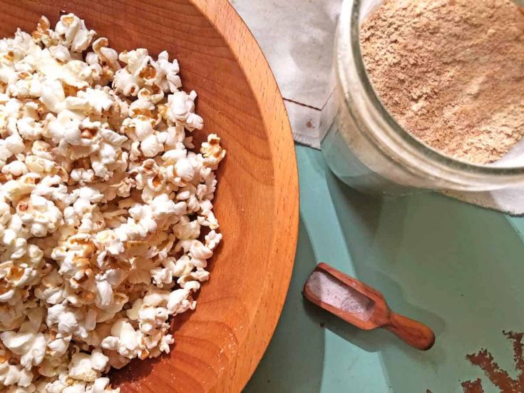 A wooden bowl is full of homemade popcorn. It is beside a glass jar of nutritional yeast and a spoonful of salt.